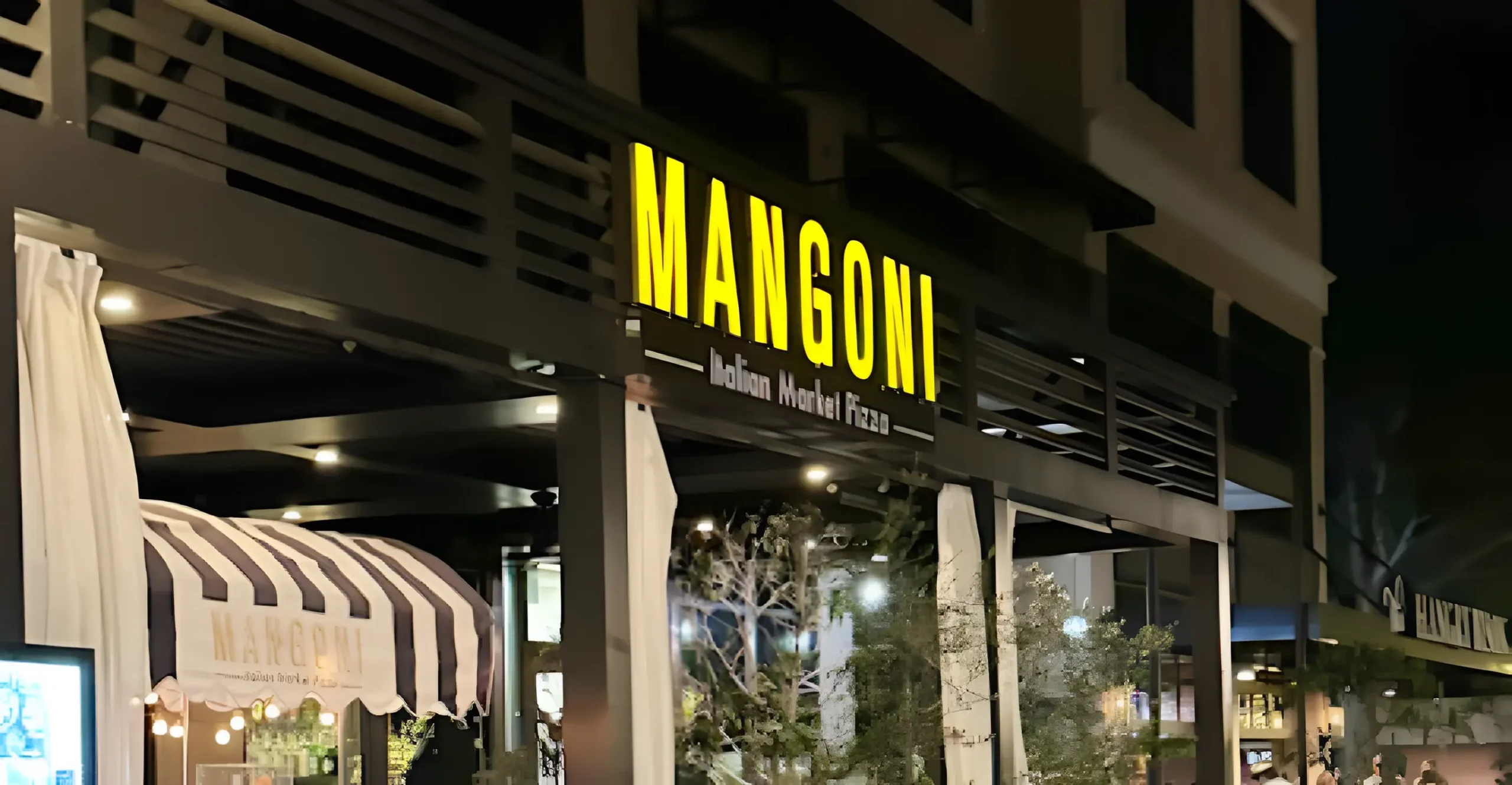 Exterior night view of Mangoni Italian Market Pizza restaurant with illuminated signage, striped awning, and cozy outdoor dining atmosphere.