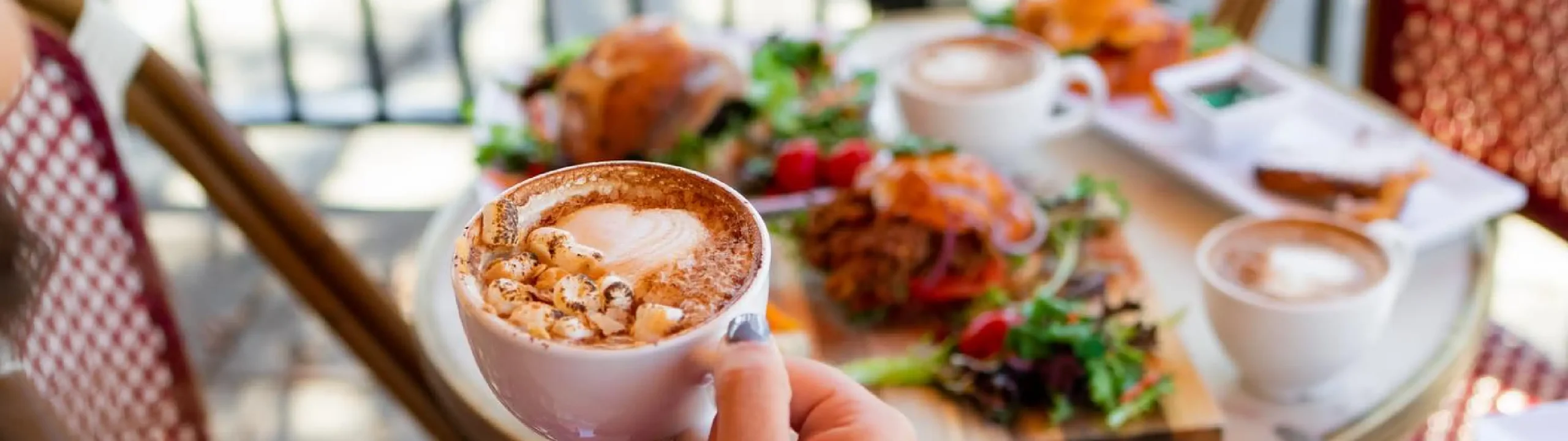 Hand holding a cup of cappuccino topped with marshmallows at Rosalie French Café in Las Vegas with brunch plates in the background