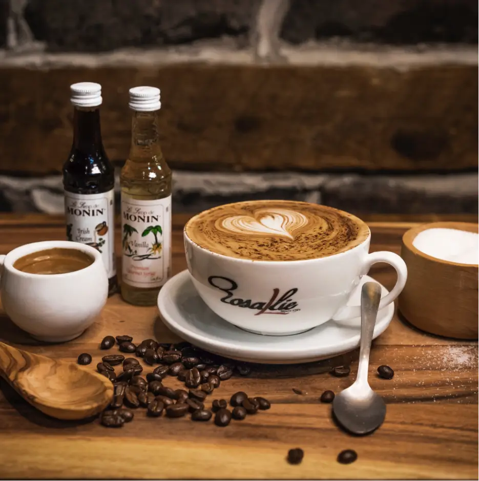 Rosallie café latte with heart-shaped foam art served alongside espresso, Monin syrups, and coffee beans on a wooden board highlighting artisanal coffee craftsmanship.